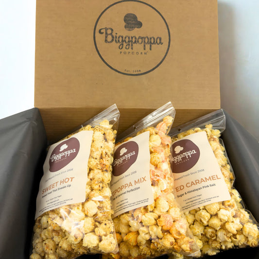 Three bags of Biggpoppa Popcorn in a rustic kraft box with visible branding.