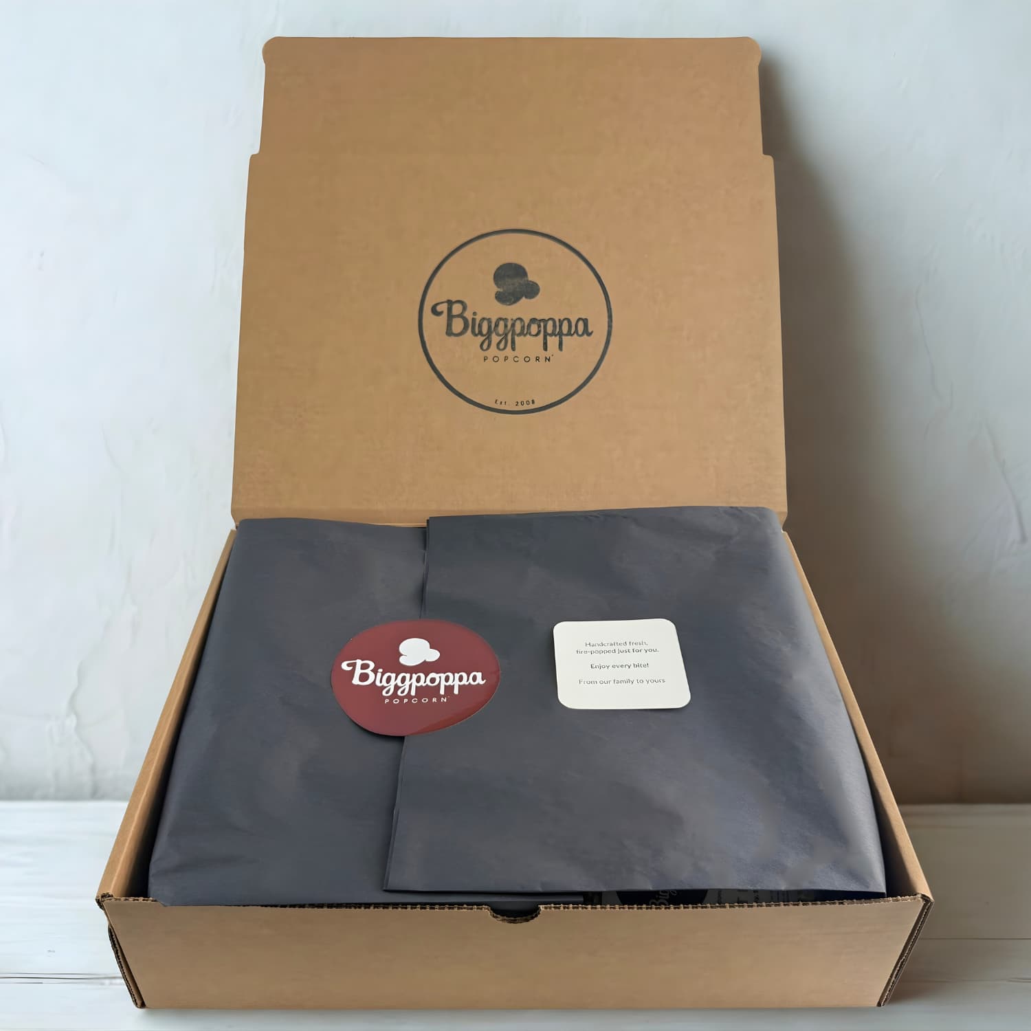 Open kraft box with popcorn wrapped in black tissue paper, sealed with a logo sticker and card.