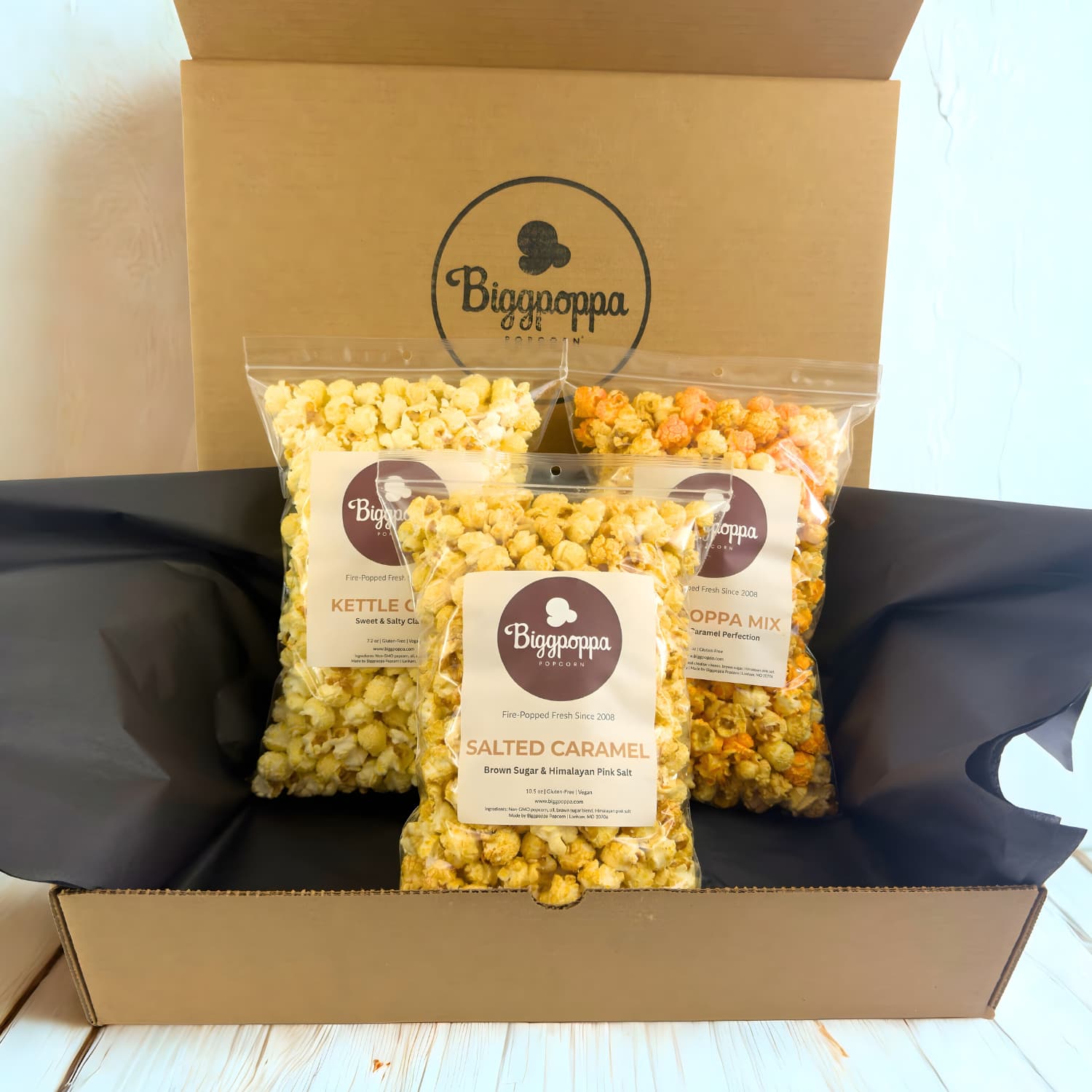 Box of Biggpoppa popcorn with three types of popcorn inside on a light wooden surface.