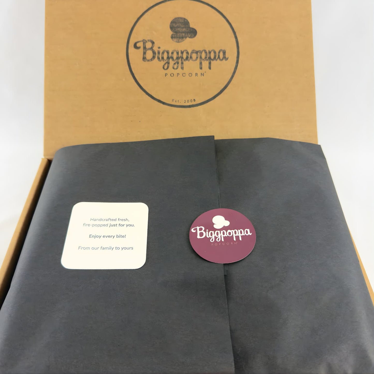 Biggpoppa popcorn packaging with box and black tissue paper, and greeting card, sealed with a sticker.